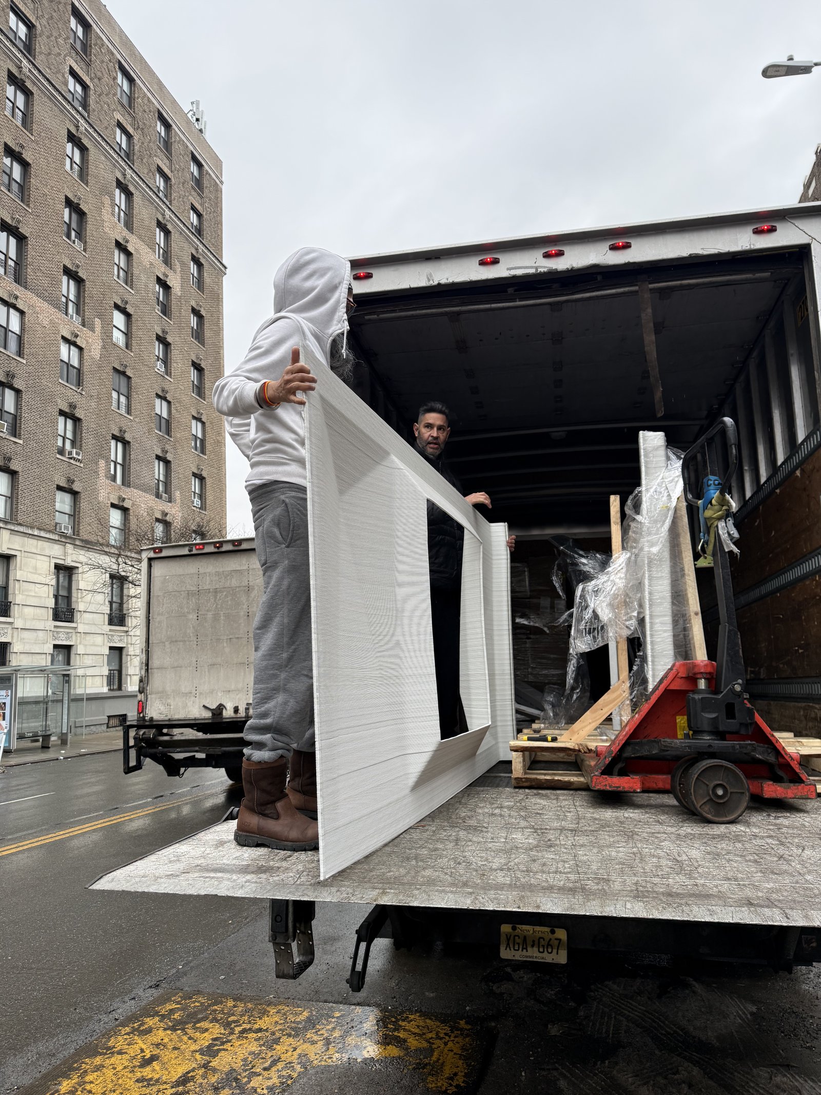 Two workers unloading an UnBOXED panel from a moving truck on a Harlem street, with pre-war apartment buildings in the background.