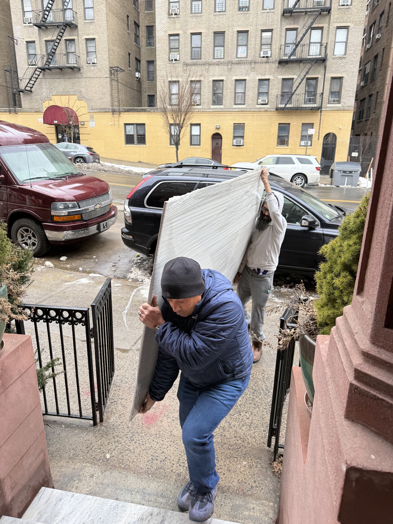 Two installers carrying a wrapped UnBOXED panel up the stoop steps of a brownstone, with apartment buildings and parked cars visible in the Harlem street behind them.