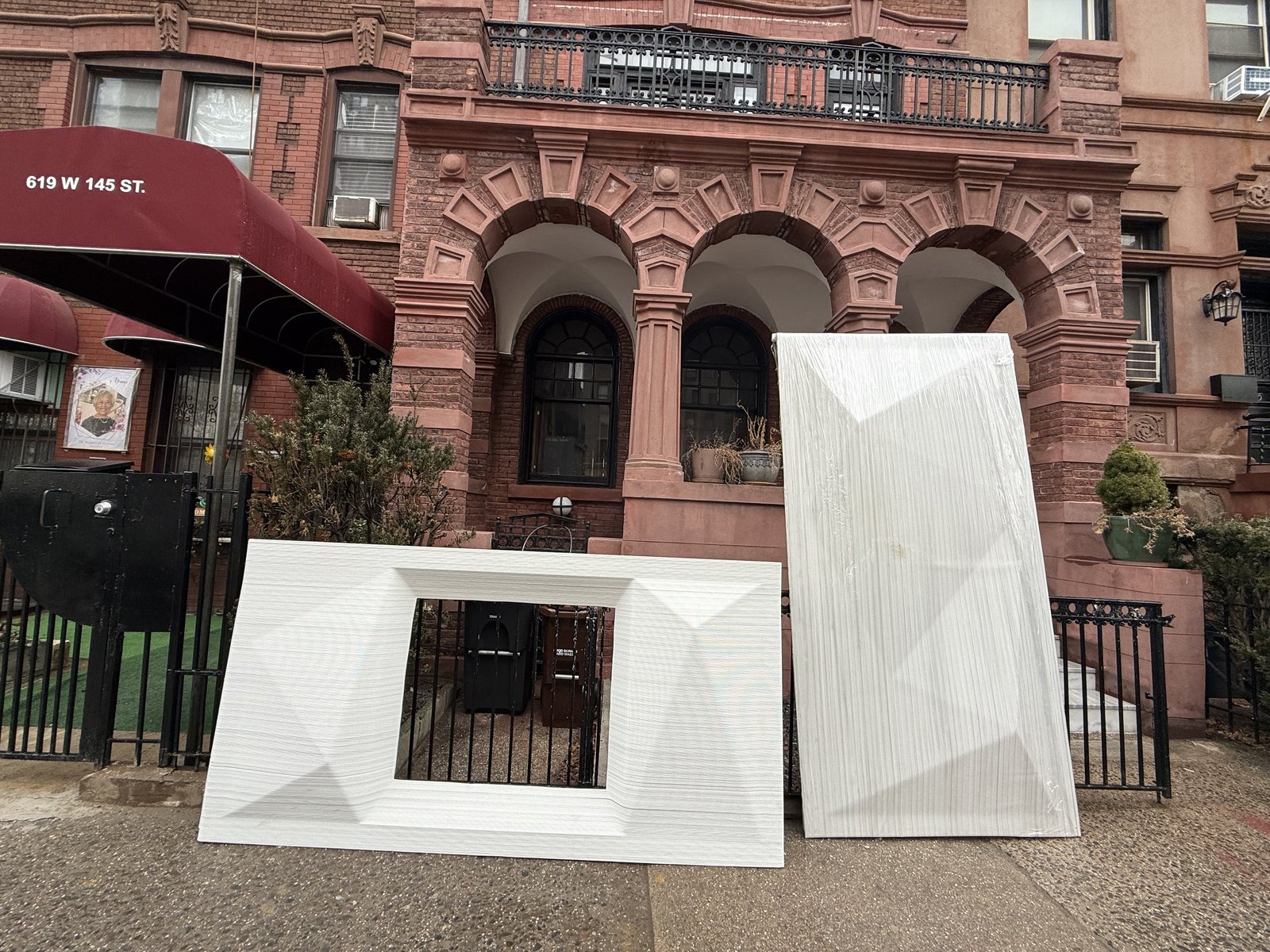 Two UnBOXED panels — one with window cutout, one wrapped — staged on the sidewalk in front of a Harlem brownstone with arched windows.