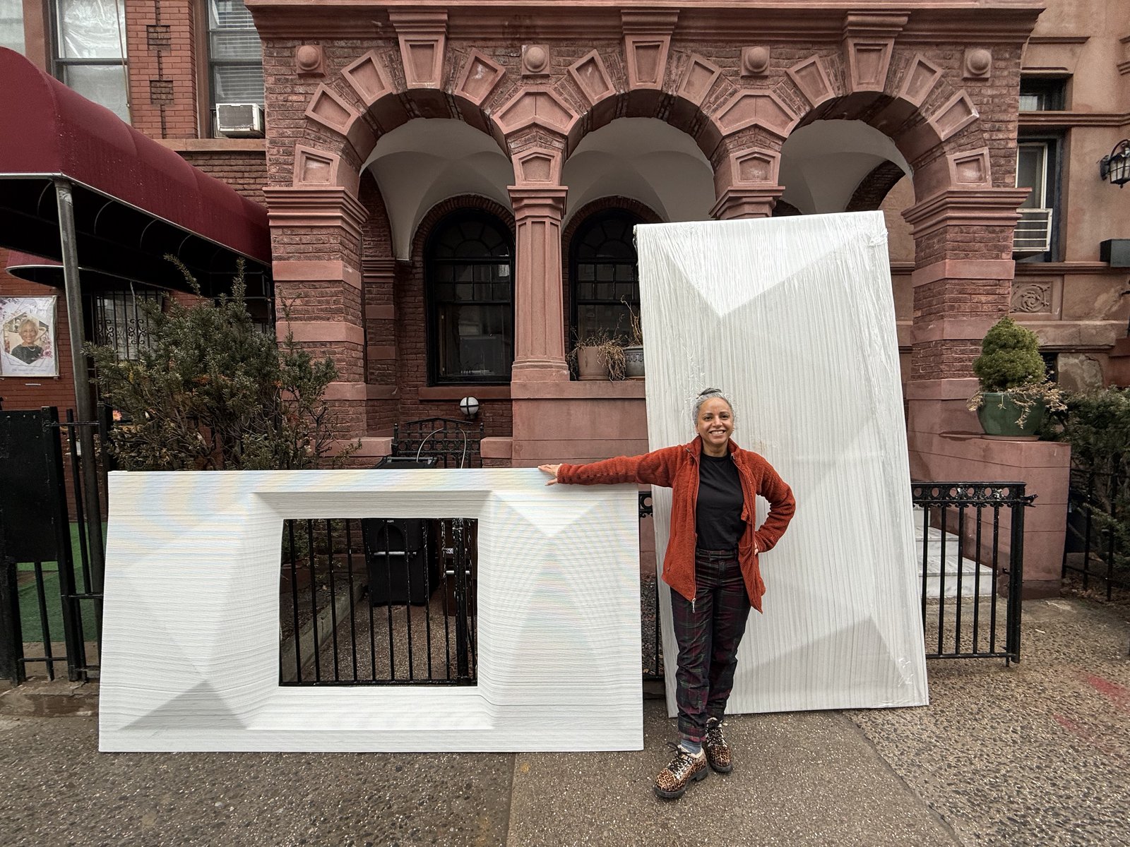 UnBOXED's founder Satpal Kaur standing between two large 3D-printed panels in front of a Harlem brownstone, smiling at the camera.